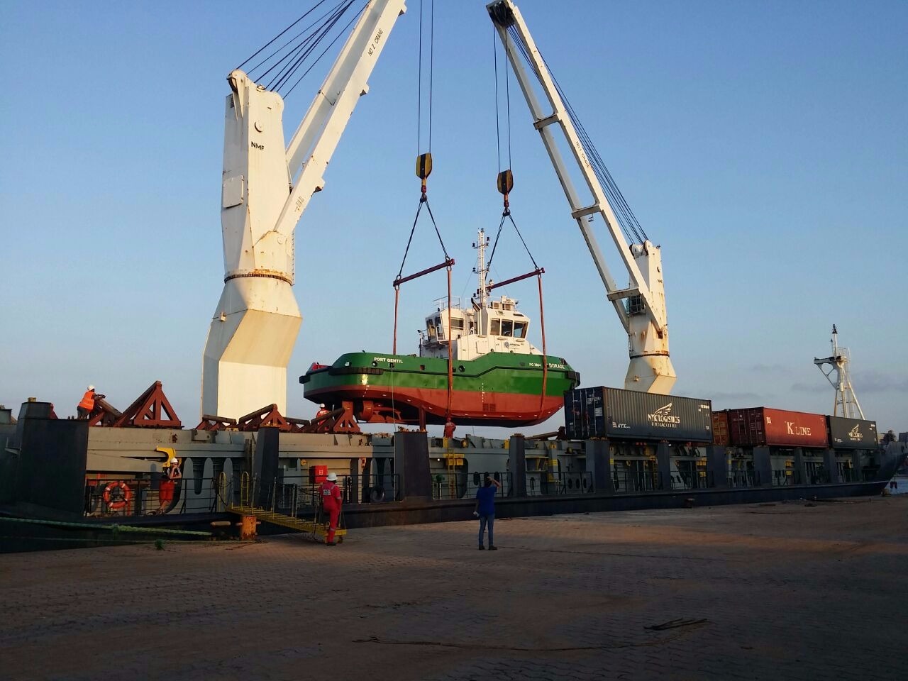 Floating crane lifting a tugboat over a barge loaded with shipping containers, with hi-vis-clad workers on site