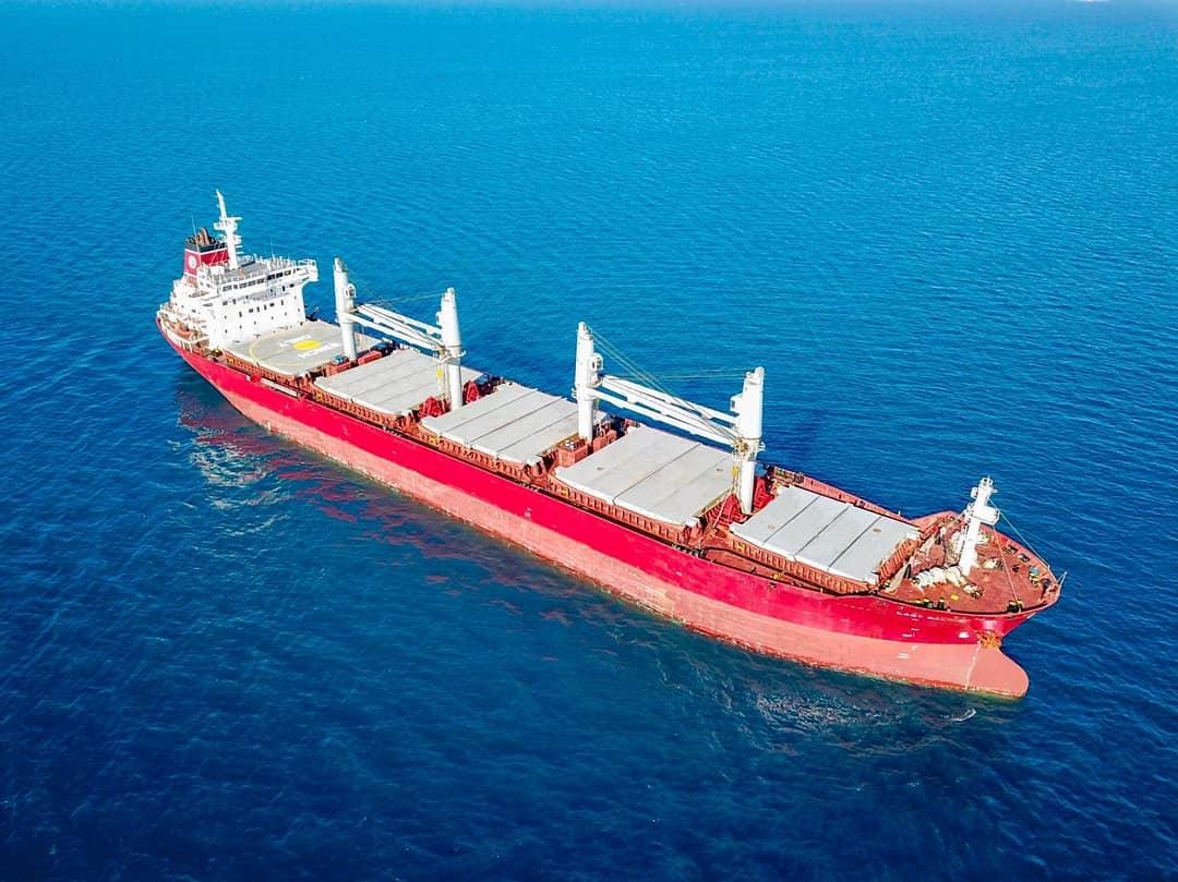 Aerial three-quarter view of a red-hulled bulk carrier at sea with open cargo hatches, sailing through deep blue water