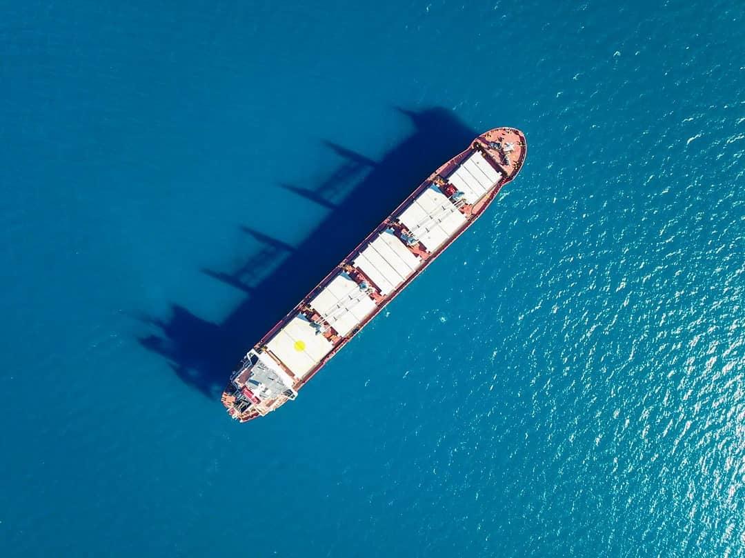 Dramatic straight-down aerial photograph of a bulk carrier casting a dark shadow on deep blue sea water
